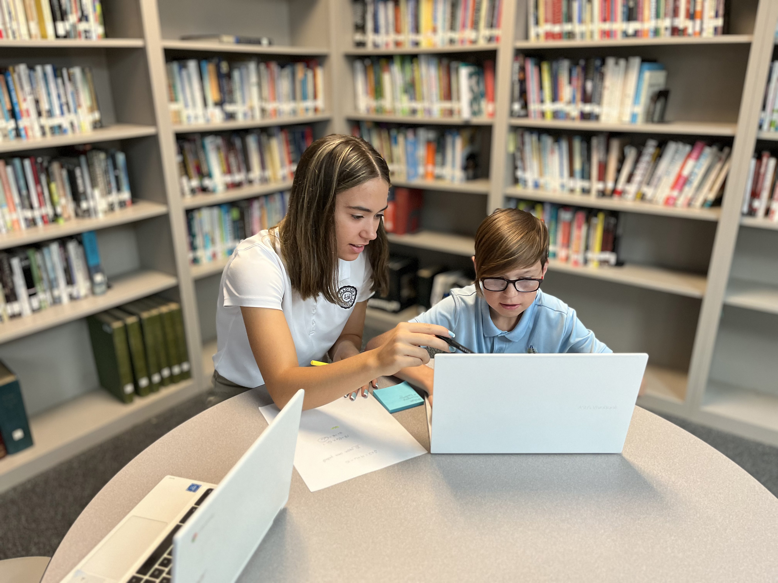 An academy student sits with an elementary student looking at a computer in a school library.